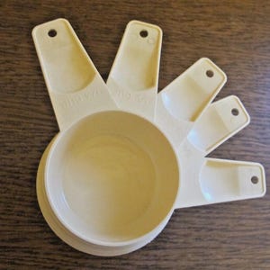 Può includere: Un set di misurini color crema, tra cui misure da 1 tazza, 1/2 tazza, 1/4 tazza e 1/8 tazza. Ogni tazza ha un manico con un foro per appenderla. Le tazze sono disposte a ventaglio su una superficie di legno.