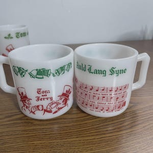 May include: White ceramic mugs with handles. One mug features green bells and the words "Tom and Jerry" with red illustrations. The other mug has "Auld Lang Syne" in green with red musical notes and lyrics.