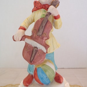 May include: A ceramic clown figurine playing a cello and holding a beach ball. The clown is wearing a yellow and red costume with a red bow tie and a white hat. The cello is brown and the beach ball is multi-colored.