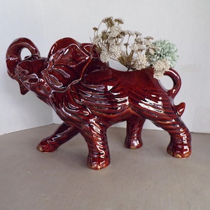 Puede incluir: Una figura de elefante de cerámica marrón brillante con la parte trasera ahuecada para sostener flores. El elefante tiene detalles y está decorado con flores secas. La figura es un artículo decorativo.