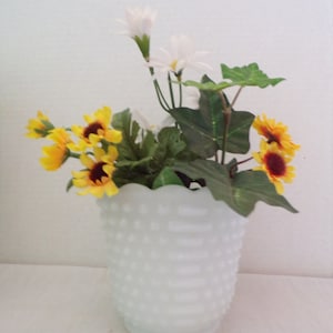 Puede incluir: Un jarrón de vidrio de leche blanco con una superficie texturizada, lleno de un ramo de flores amarillas y blancas. Las flores incluyen margaritas y girasoles.