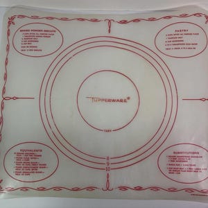 Op de afbeelding: Een doorschijnende, vierkante Tupperware bakmat met rode bedrukte tekst en afbeeldingen. De mat bevat concentrische cirkels voor taartmaten, gelabeld met 8, 9 en 10 inch, samen met recepten voor biscuits, gebakrecepten, equivalenten en vervangingen.