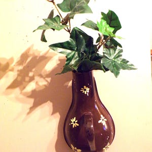 Puede incluir: Un jarrón de cerámica marrón con una silueta en forma de pera y diseños florales blancos. El jarrón está lleno de plantas verdes.