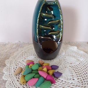 May include: A teal and black ceramic incense waterfall with a cascading design. A collection of colorful incense cones in shades of pink, green, yellow, and purple are in front of the waterfall.
