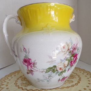 May include: A large, white ceramic vase with a yellow top and floral design. The vase has two handles and features a pattern of pink and white roses with green leaves.