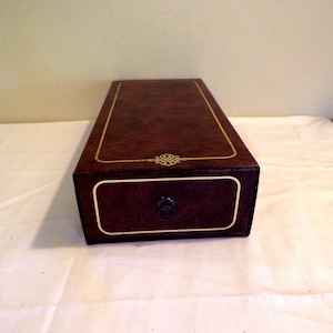 May include: A rectangular, dark brown box with a drawer, featuring gold trim and a decorative emblem. The drawer has a small, round, black handle. The box is made of a textured material.