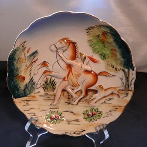 Puede incluir: Plato decorativo de cerámica con borde festoneado. El plato presenta una escena en relieve de una persona a caballo, con un perro corriendo al lado. La paleta de colores incluye tonos de marrón, naranja, verde y azul.