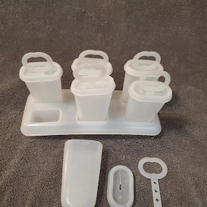 Puede incluir: Un juego de moldes de plástico blanco para helados. El juego incluye una bandeja con seis moldes individuales, un molde más grande, una tapa y una pequeña herramienta. Los moldes tienen asas. Los artículos están sobre una superficie gris.