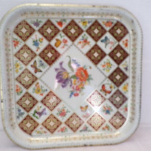 Puede incluir: Una bandeja de servir de metal blanco con un borde dorado y un patrón floral. El patrón presenta un diseño de diamante repetido con acentos rojos, marrones y dorados. El centro de la bandeja presenta un ramo de flores con mariposas.