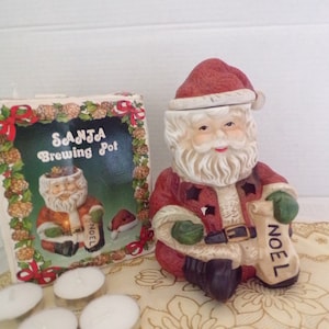May include: A ceramic Santa Claus figurine holding a sack that says "Noel". The figurine is red and white with a black belt. The figurine is a wax warmer and has a hole in the top for a tea light candle. The box says "Santa Brewing Pot".