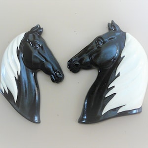 Op de afbeelding: Twee zwarte en witte keramische wanddecoraties van paardenhoofden. De paarden hebben witte manen en staarten. De decoraties zijn naar elkaar gekeerd.