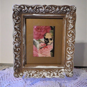 May include: A white and brown ornate picture frame with a skull and flowers image inside. The frame has a distressed white paint finish and intricate scrollwork.