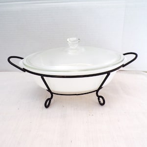 May include: White oval glass baking dish with a glass lid, sitting on a black metal stand with three legs.
