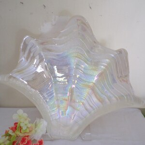 May include: Iridescent glass bowl with a wavy, textured design. The bowl has a star-like shape with a wide, open center. The glass reflects a rainbow of colors. A small bouquet of flowers is in the foreground.