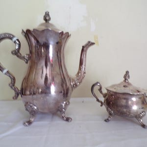 Op de afbeelding: Een zilverkleurige theeset, bestaande uit een hoge theepot met een gebogen tuit en sierlijke handgreep, en een kleinere suikerpot met deksel. De set heeft decoratieve details en staat op een wit oppervlak.