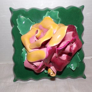 May include: A ceramic wall plaque featuring a three-dimensional rose with yellow, pink, and burgundy petals. The rose is set against a green background with leaves.