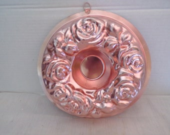 Puede incluir: Un molde de pastel bundt de cobre con un diseño de rosas. El molde circular tiene un agujero central y un patrón decorativo de rosas alrededor del borde. Se adjunta un pequeño lazo para colgar.