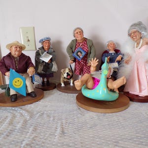 May include: A collection of whimsical figurines depicting elderly people in various poses. One figure holds a blue flag with a smiley face, while another is in a pool float. Other figures include a dog and a woman in a pink dress.