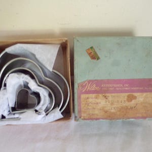 Puede incluir: Juego de cortadores de galletas en forma de corazón vintage en una caja de cartón. El juego incluye cortadores de metal de varios tamaños anidados. La caja está abierta, revelando los cortadores y papel de seda.