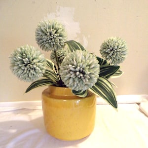 May include: A yellow ceramic vase holds a floral arrangement. The arrangement features several spherical, light green flowers and green and white striped leaves. The vase is a solid, bright yellow color.
