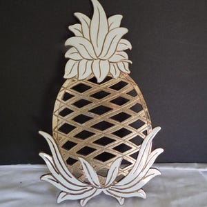 Puede incluir: Un objeto decorativo en forma de piña con un cuerpo dorado y detalles blancos. La piña tiene un diseño de diamantes y hojas blancas en la base y en la parte superior. El objeto probablemente está hecho de madera o metal.