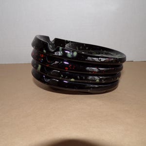 Puede incluir: Cenicero negro con un diseño circular apilado. El cenicero tiene un acabado brillante con detalles moteados rojos, verdes y blancos. El cenicero tiene una ranura para apoyar los cigarrillos.