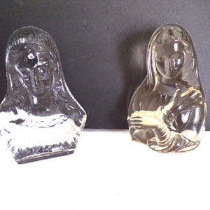 May include: Two clear glass sculptures of women, one with her arms crossed and the other with her hands clasped in front of her.