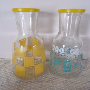 May include: Two vintage glass pitchers with yellow lids. One pitcher has a yellow and white checkered pattern, while the other has a teal and white design. Both pitchers have a ribbed neck and a bulbous body.