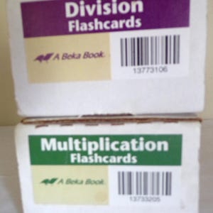May include: Two white boxes of educational flashcards. The top box has a purple banner with "Division Flashcards" in white text. The bottom box has a green banner with "Multiplication Flashcards". Both boxes have a barcode and "A Beka Book" logo.