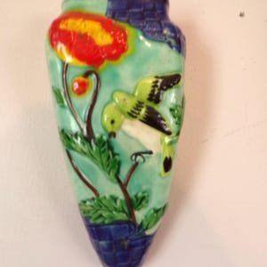May include: A ceramic wall pocket vase with a colorful floral and bird design. The vase features a red and yellow flower, a green bird, and green leaves against a turquoise background. The top and bottom of the vase are blue.