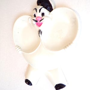 Puede incluir: Decoración de pared de cerámica blanca con un personaje de dibujos animados sosteniendo dos cuencos. El personaje tiene un sombrero negro, ojos negros, una nariz rosa y una boca rosa.