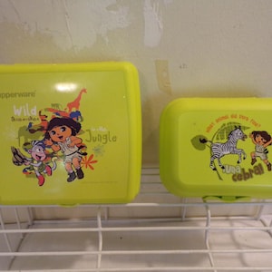 May include: Two yellow Tupperware lunchboxes with cartoon graphics. The larger box features Dora the Explorer with a monkey and the words "Wild in the Jungle." The smaller box has a zebra and Dora with the words "Una cebra."
