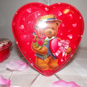 May include: Red heart-shaped tin box featuring a teddy bear holding a bouquet of red roses and a heart-shaped box with a pink bow. The tin is decorated with white hearts.