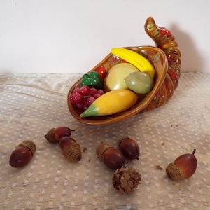 Puede incluir: Una cornucopia de cerámica rebosante de coloridas frutas artificiales, incluyendo un plátano, uvas y un mango. Varias bellotas están esparcidas alrededor de la base del cuerno de la abundancia. La cornucopia es marrón y naranja.