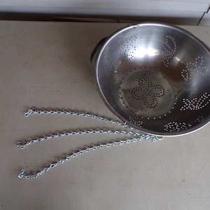 Puede incluir: Un colador de acero inoxidable con perforaciones decorativas y dos asas. Se adjuntan tres cadenas plateadas, probablemente para colgar. El colador está diseñado para escurrir líquidos de los alimentos.