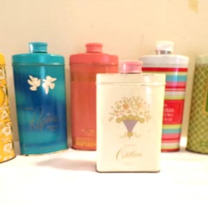 Op de afbeelding: Een verzameling vintage metalen containers in verschillende kleuren, waaronder geel, blauw, rood en groen. Sommige containers hebben bloemmotieven en tekst. De containers hebben deksels en zijn gerangschikt op een wit oppervlak.