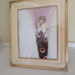 Pode incluir: Uma impressão de arte emoldurada com uma ilustração estilizada de uma mulher em um vestido preto com padrões circulares vermelhos e amarelos. A obra de arte está em um fundo rosa e emoldurada em uma moldura creme com detalhes dourados nos cantos.