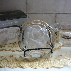 May include: A clear glass trinket dish with a rose design. The dish is on a black metal stand and is surrounded by a gold lace doily. The dish is packaged in a box that says "Crystal Jewelry Trinket Dish".