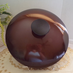 Könnte beinhalten: Ein brauner Deckel für einen Topf oder eine Pfanne mit einem schwarzen Knopf oben. Der Deckel liegt auf einer weißen Spitze.