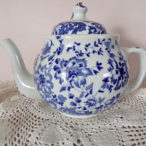 Könnte beinhalten: Weiße Keramik-Teekanne mit blauem Blumenmuster. Die Teekanne hat einen gebogenen Ausguss, einen Henkel und einen Deckel mit einem kleinen Knopf. Das blaue Blumendesign bedeckt die gesamte Oberfläche der Teekanne.