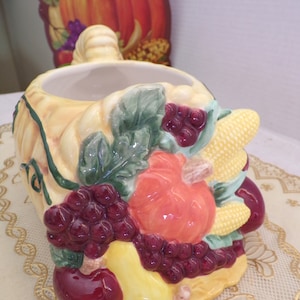 Puede incluir: Un cuenco de cerámica con forma de cornucopia decorado con una variedad de frutas y verduras de otoño, incluyendo uvas, una pera, una manzana, una calabaza y maíz. El cuenco está pintado en colores realistas y tiene un estilo rústico, de granja.