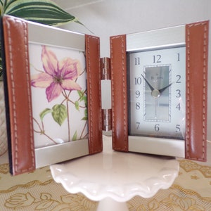 May include: A brown leather-bound picture frame with a silver metal frame and a floral print inside. The frame is open to reveal a silver clock face with black hands and Roman numerals.