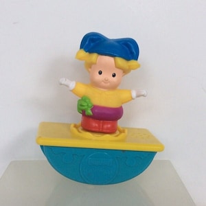 May include: A yellow and blue toy seesaw with a small, yellow-clad figure with a blue hat and blonde hair standing on top. The figure is holding a green bow. The toy is labeled "Little People".