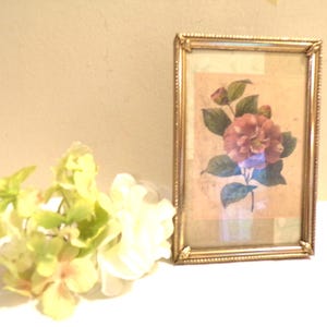 May include: A framed floral print featuring a pink flower with green leaves. The frame is gold-colored with a decorative edge. A small bouquet of light green and white flowers is in the foreground, creating a vintage aesthetic.