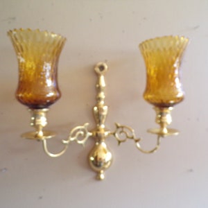 May include: A brass wall sconce with two amber glass candle holders. The sconce has a decorative scroll design.