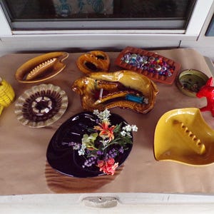 以下が含まれることがあります： 黄色、茶色、黒など、さまざまな形と色のヴィンテージセラミック灰皿のコレクション。装飾的な要素を持つ灰皿もあり、小さな赤い置物も特徴です。いくつかの灰皿には小さなオブジェクトが含まれています。