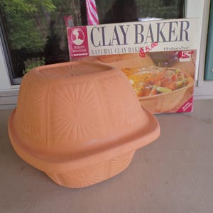 Op de afbeelding: Een terracotta kleibakvorm met een deksel voorzien van een zonnestraalpatroon aan elke kant. De productdoos is gelabeld met CLAY BAKER NATURAL CLAY BAKER en vermeldt afmetingen van 33 x 24,8 x 19 cm, met het Baker's Advantage by Rosco logo zichtbaar.