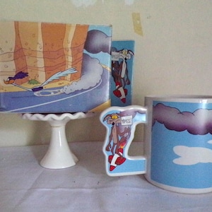 May include: A ceramic mug and a decorative box featuring cartoon characters. The mug has a blue sky design with a road sign. The box shows a roadrunner speeding down a road. The handle of the mug has a cartoon character design.