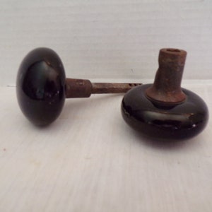 Może przedstawiać: Dwa antyczne czarne ceramiczne gałki drzwiowe z metalowymi trzpieniami. Gałki są okrągłe i mają gładkie wykończenie.