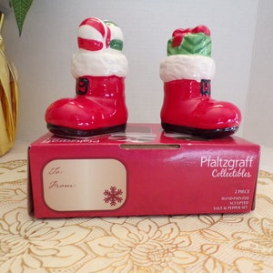 Op de afbeelding: Een zout- en peperstrooierset in de vorm van kerstmansschoenen, rood en wit. De schoenen zijn versierd met snoepstokken, cadeautjes en witte bont. De doos heeft de tekst "Pfaltzgraff Collectibles" en "2-delige handgeschilderde, gebeeldhouwde zout- en peperset".